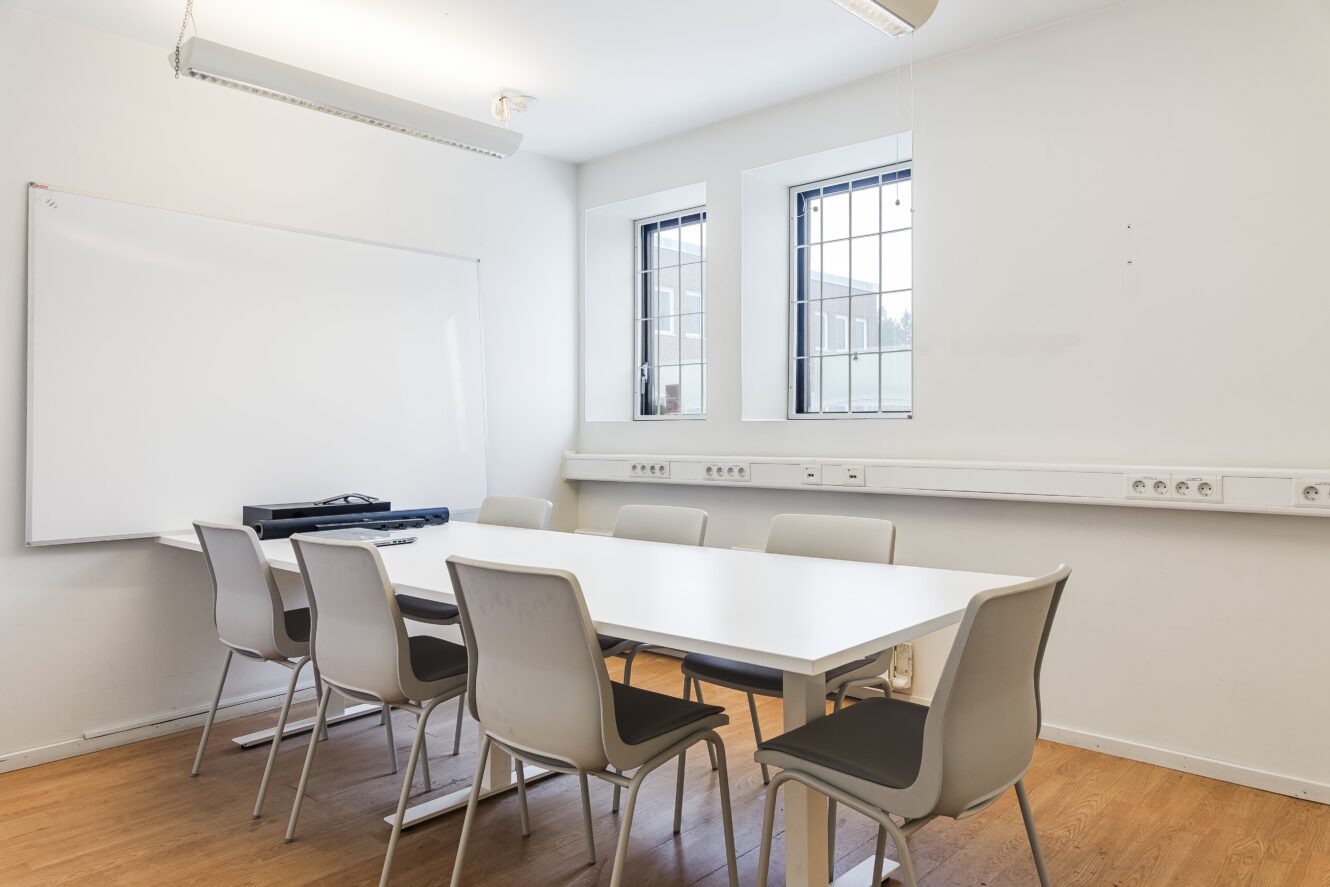 Ett litet konferensrum med ett vitt bord, åtta grå stolar, en whiteboard och två fönster med galler, beläget på ett trägolv.