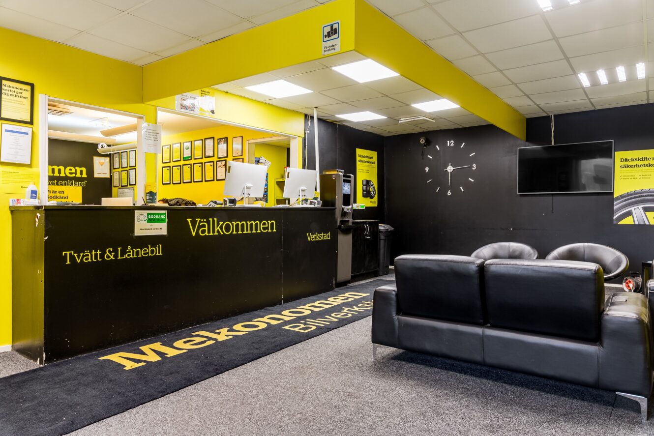 Receptionsområdet på en bilverkstad med gul och svart färgsättning, en disk med datorer, en väggklocka, en svart soffa och informationsskyltar på väggarna.