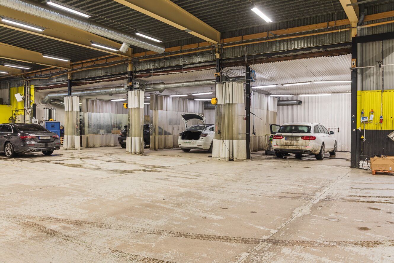 Tre bilar står parkerade inne i en bilverkstad med öppna arbetsplatser och takbelysning; en av bilarna har bagageluckan öppen.