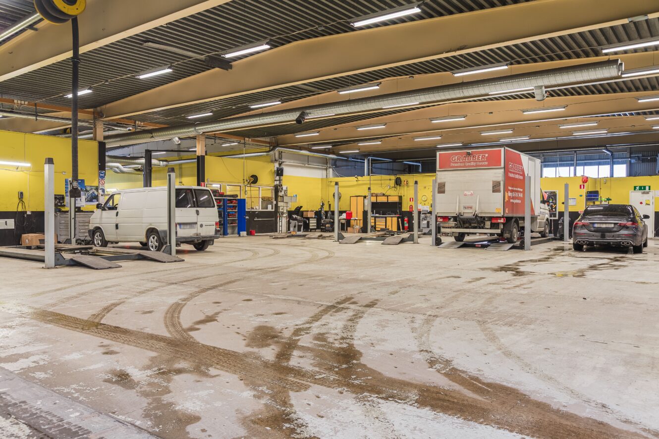 En rymlig bilverkstad med gula väggar, flera fordon – däribland en skåpbil och en lastbil – på lyftbryggor, verktyg och utrustning uppställda längs väggarna samt däckspår på golvet.