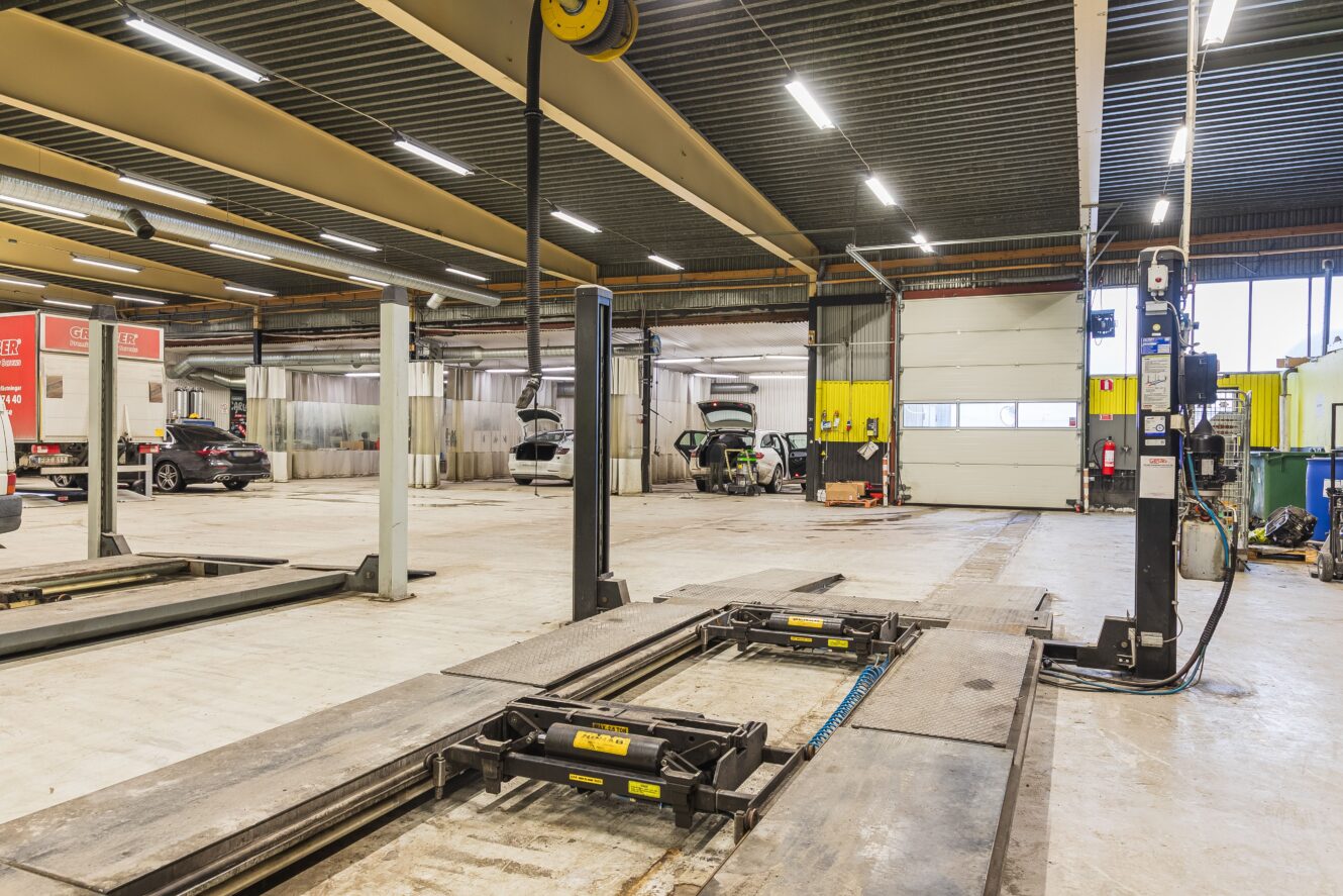 En vid vy över en rymlig bilverkstad med billyftar, fordon som håller på att servas och diverse utrustning utspridd i den rena och välorganiserade verkstaden.