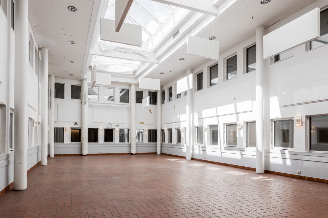 Rymligt, tomt atrium med stora fönster, vita väggar, takfönster och rött klinkergolv. Naturligt ljus fyller området från ovan.