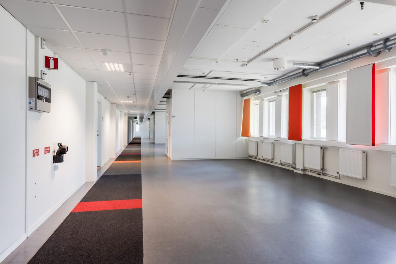 En modern, tom korridor med vita väggar, stora fönster till höger och ett grått golv med röda och svarta mattor.