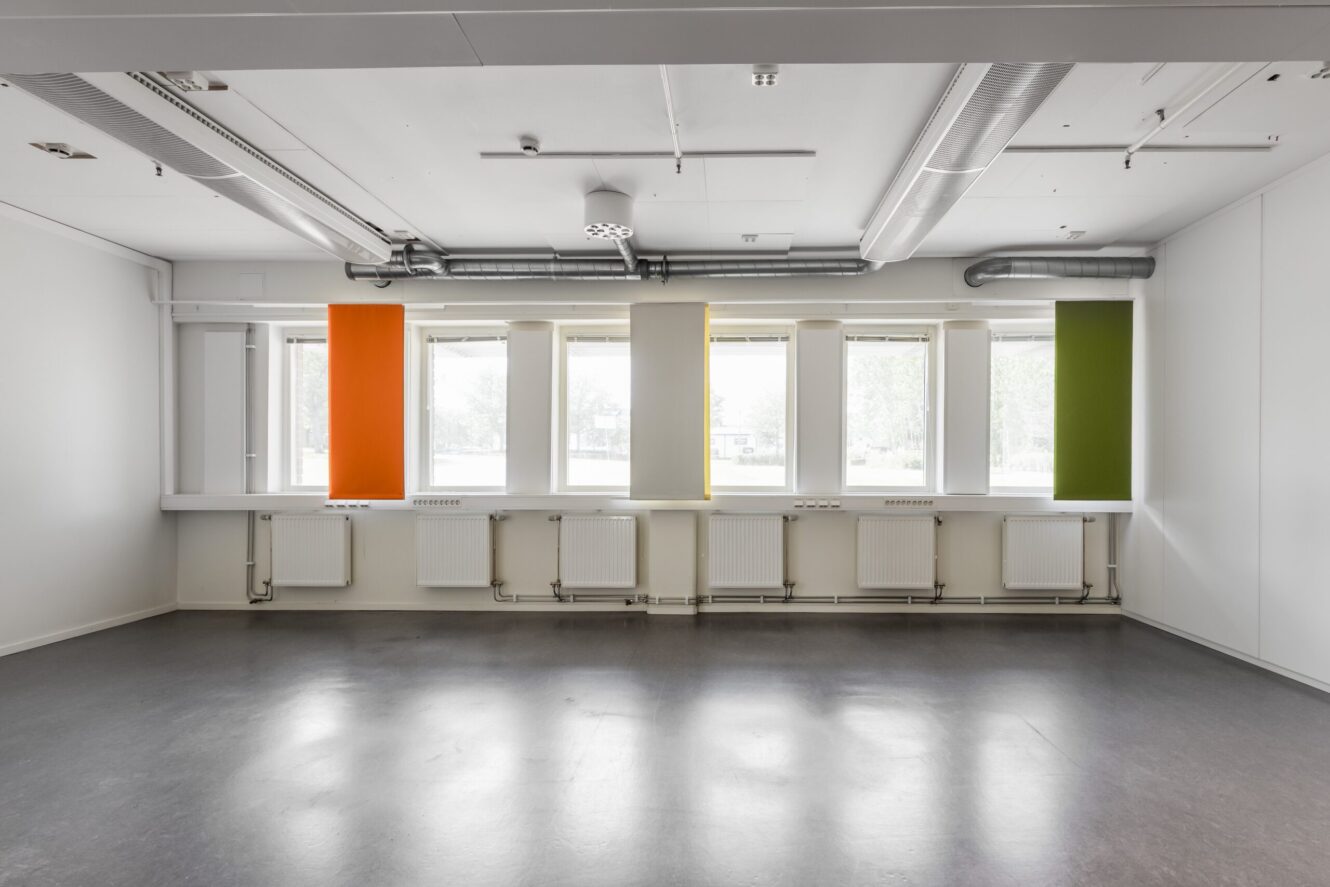 Tomt rum med vita väggar, grått golv, stora fönster och tre vertikala paneler i orange, gult och grönt som hänger framför fönstren.