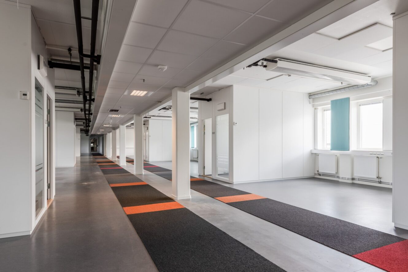 En modern, tom hall med grått golv, vita väggar, stora fönster och mönstrade mattor i svart, grått och orange. Rör i taket och öppna dörröppningar är synliga.