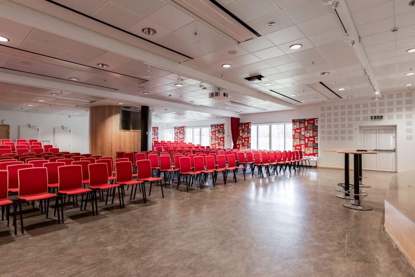 Ett stort konferensrum med rader av tomma röda stolar som vetter mot en scen, bord och fönster längs bakväggen.