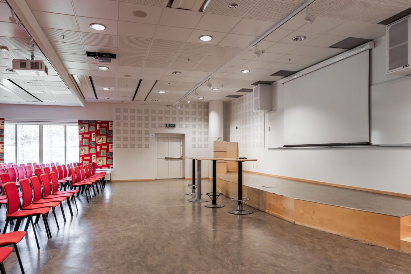 Tom föreläsningssal med rader av röda stolar, ett podium i trä på en scen, runda bord och en stor projektorduk längst fram.