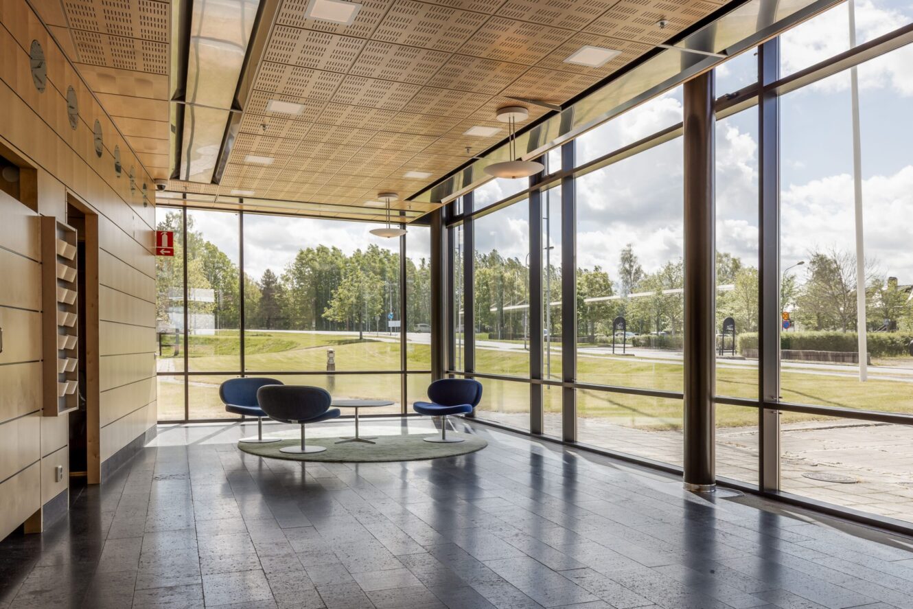 Modern interiör med stora fönster från golv till tak, tre runda stolar runt ett lågt bord på en matta och utsikt över träd och gräs utanför.