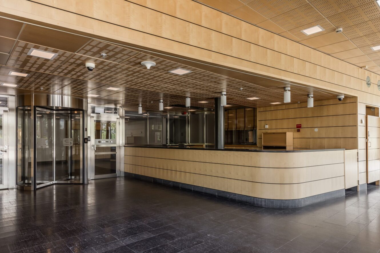 Lobby i modern byggnad med väggpaneler i ljust trä, en stor tom receptionsdisk, svart golv och en karuselldörr i glas till vänster.