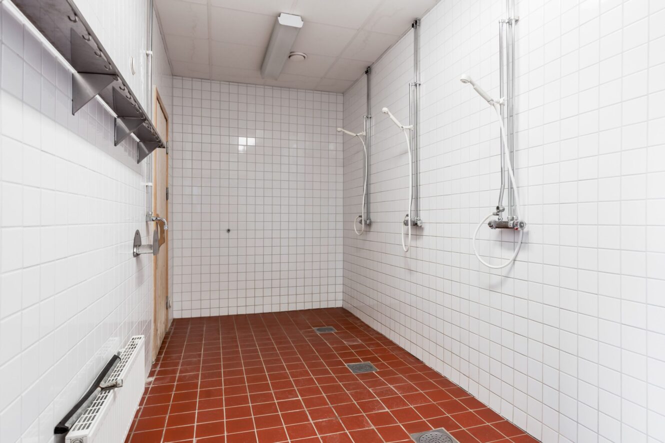 Ett vitkaklat gemensamt duschrum med tre duschmunstycken, rött klinkergolv, metallhyllor och en trädörr.