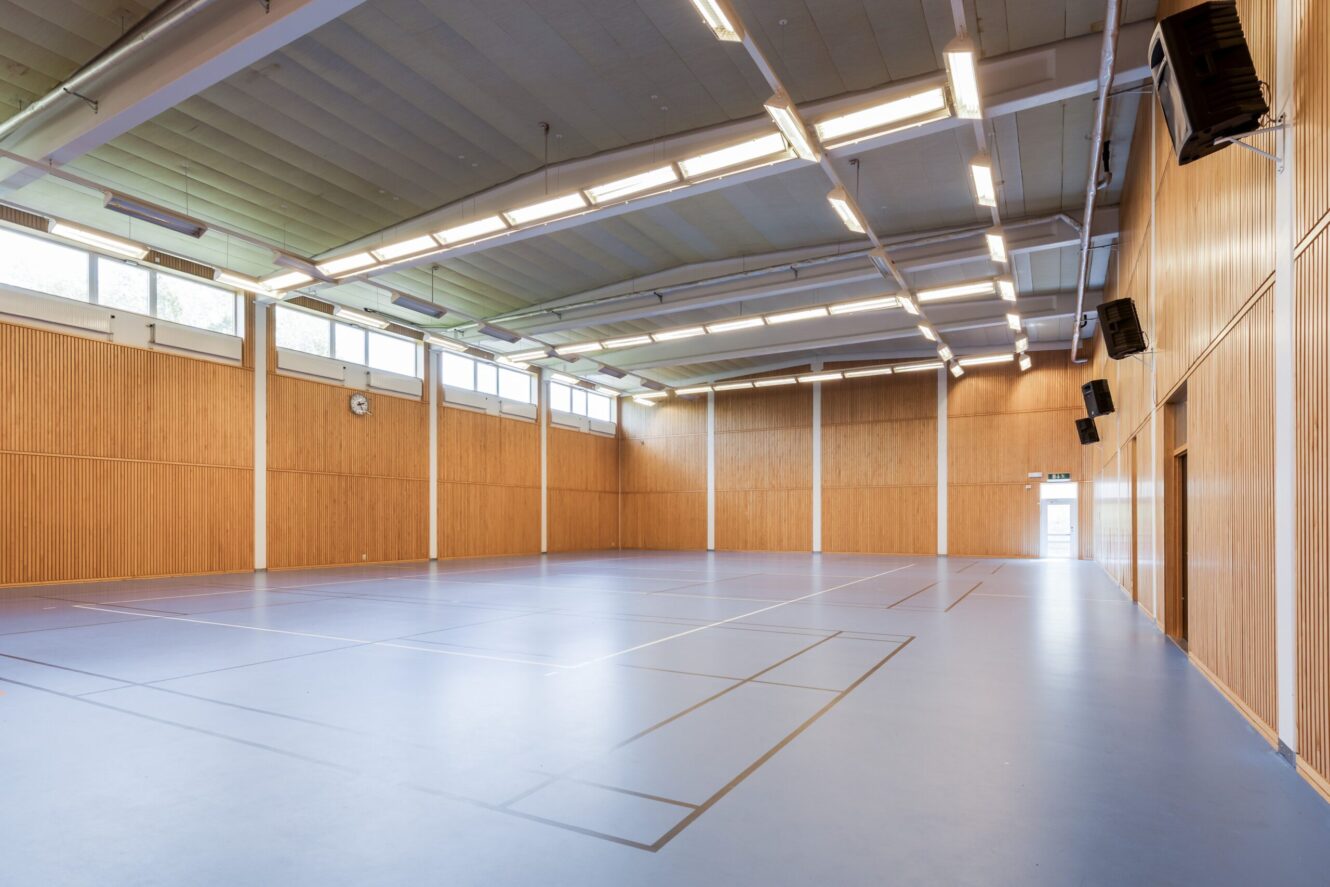 Stor, tom gymnastiksal inomhus med ljusblått golv, markerade sportlinjer, träpanel på väggarna, högt i tak och fönster längs de övre väggarna som släpper in naturligt ljus.