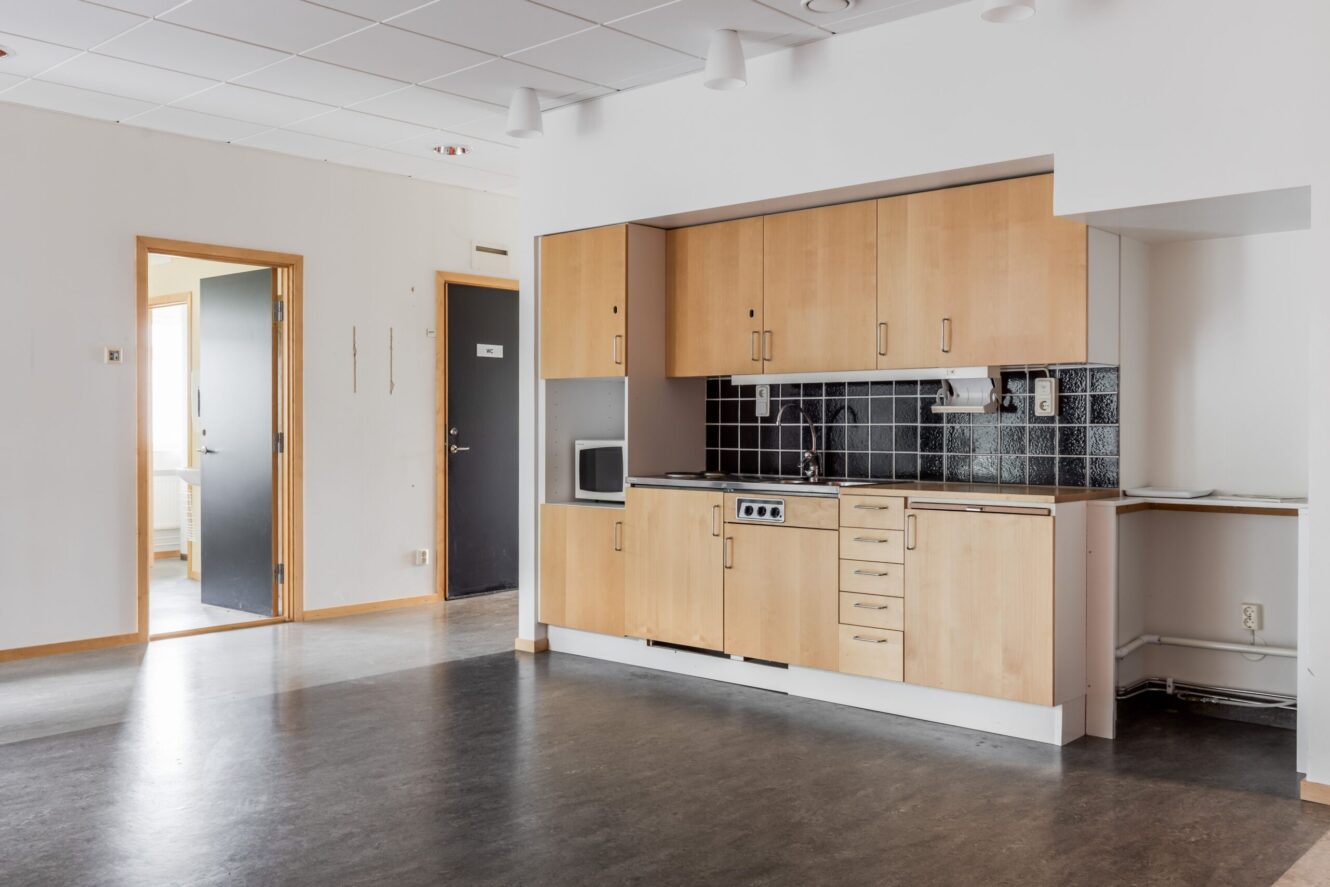 Omöblerad lägenhet med ett litet kök med skåp i ljust trä, svart kakel och öppna dörrar som leder till andra rum.