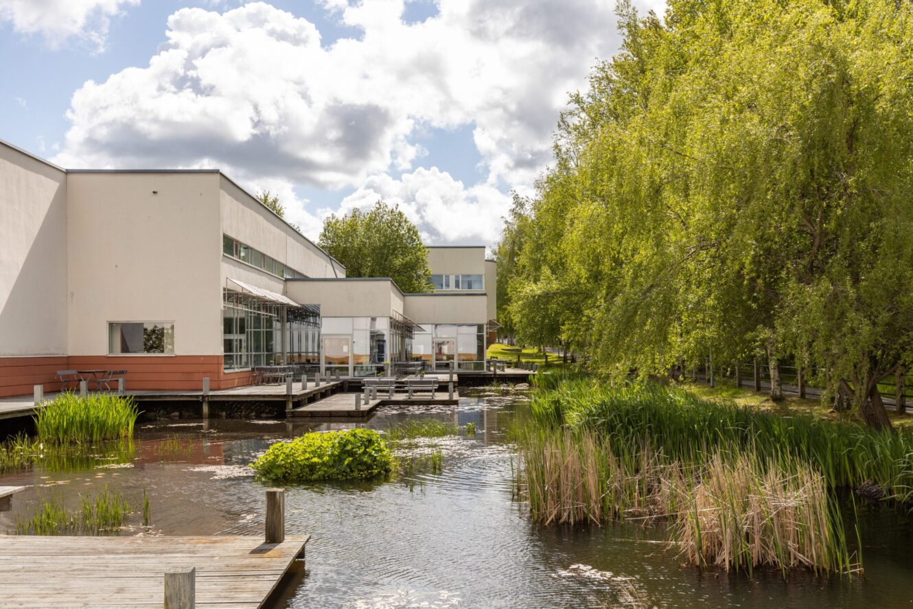 Modern vit byggnad med stora fönster intill en damm, omgiven av grönskande träd och vass under en delvis molnig himmel.