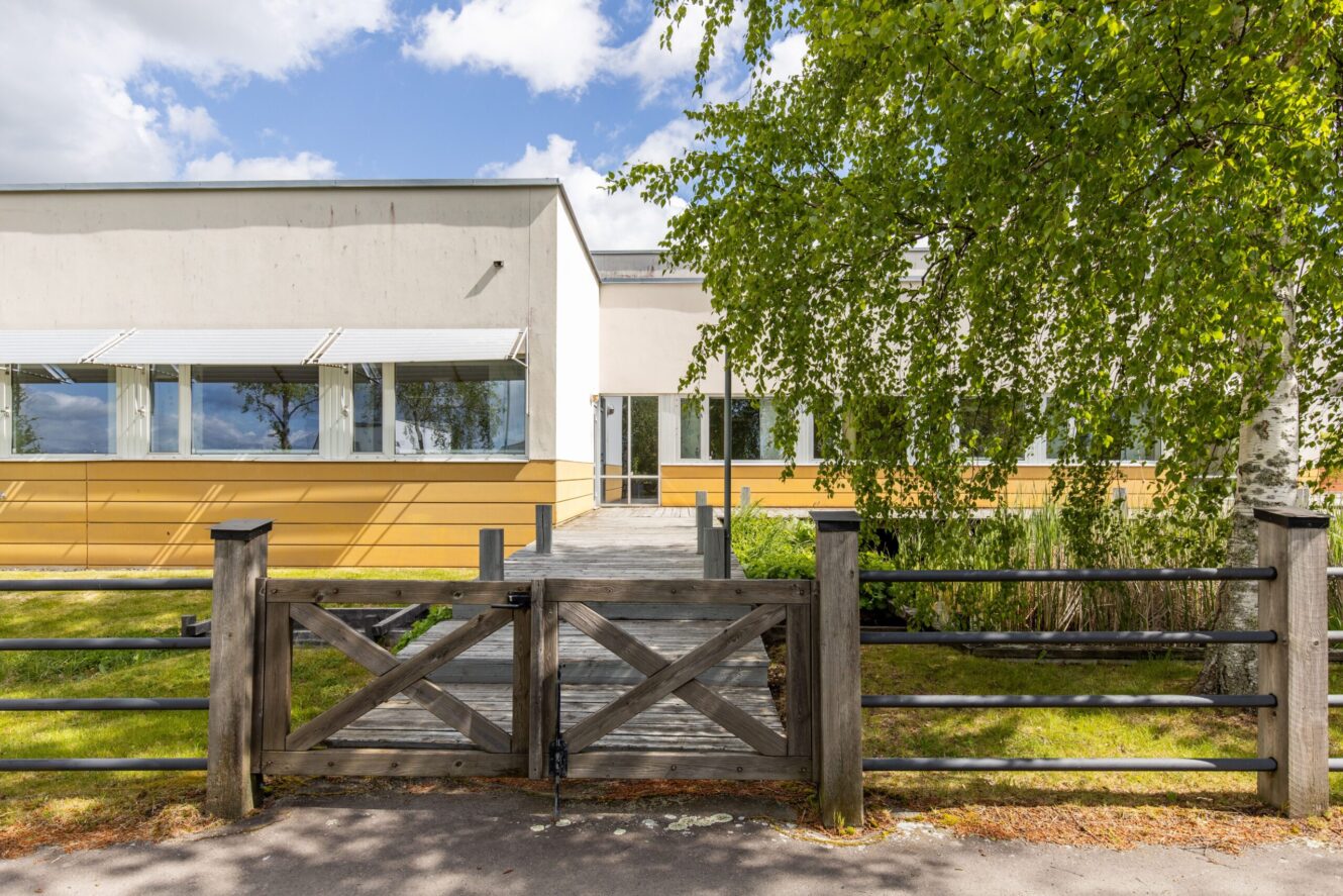 Envåningsbyggnad med stora fönster och gul nedre fasad, sedd bakom en grind och ett staket av trä, med träd och grönska i förgrunden.