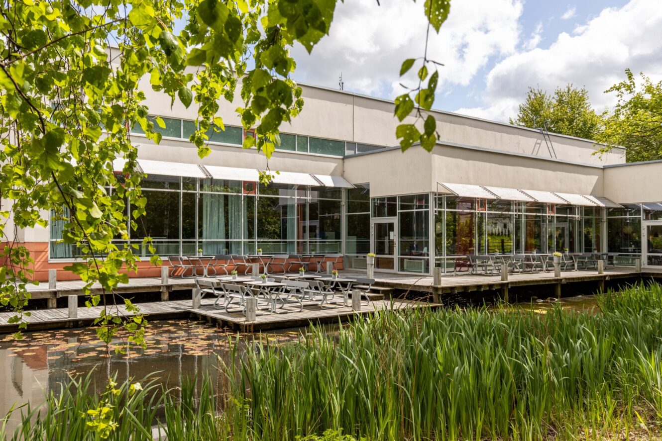 Modern byggnad med stora fönster och uteservering på ett trädäck med utsikt över en damm med vattenväxter och lövträd i förgrunden.