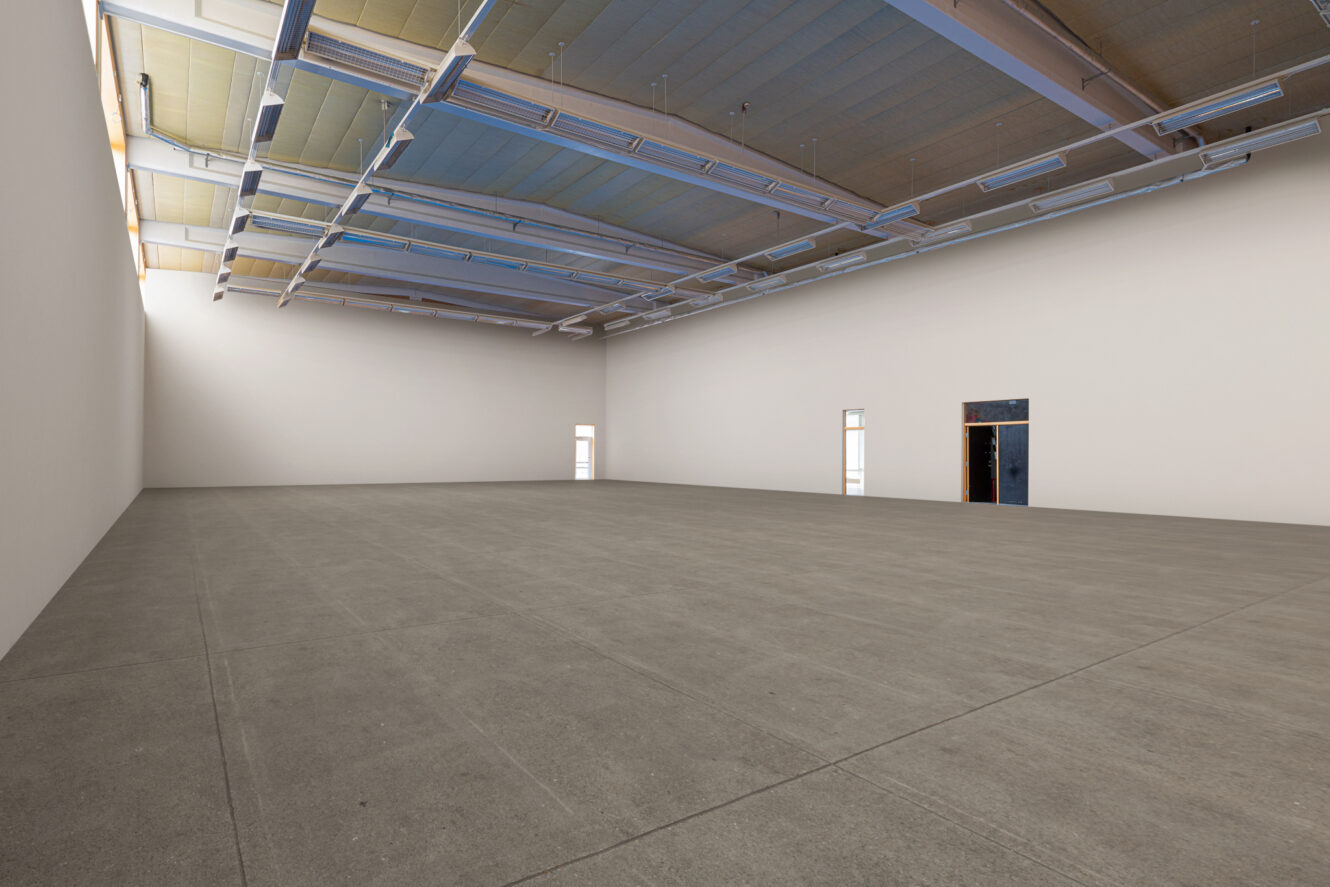 Stort tomt rum med högt i tak, vita väggar, betonggolv och flera små dörrar längs den bortre väggen.