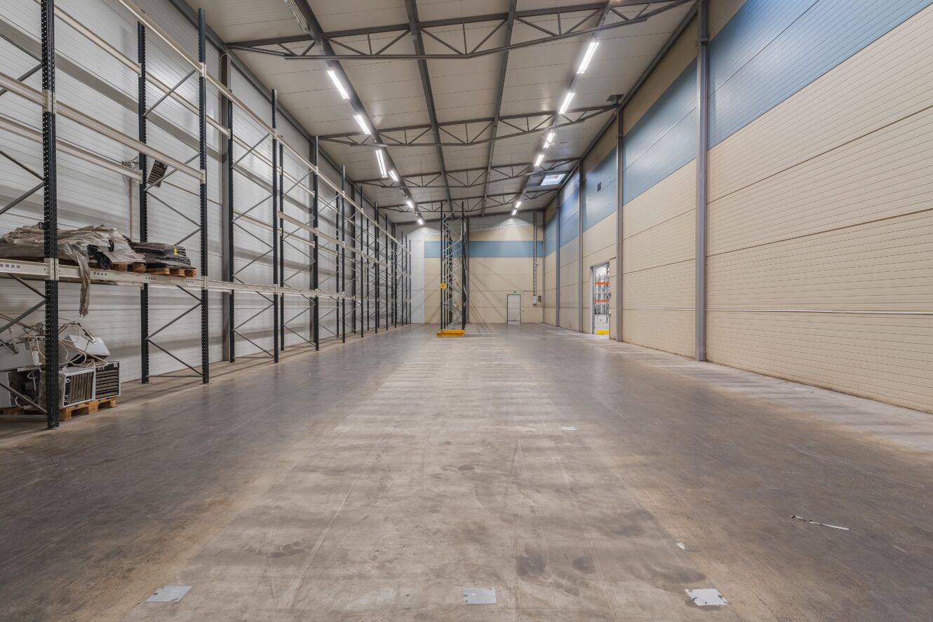 Tomt industrilager med högt i tak, synliga bjälkar, metallhyllor till vänster och ett rent betonggolv under stark takbelysning.