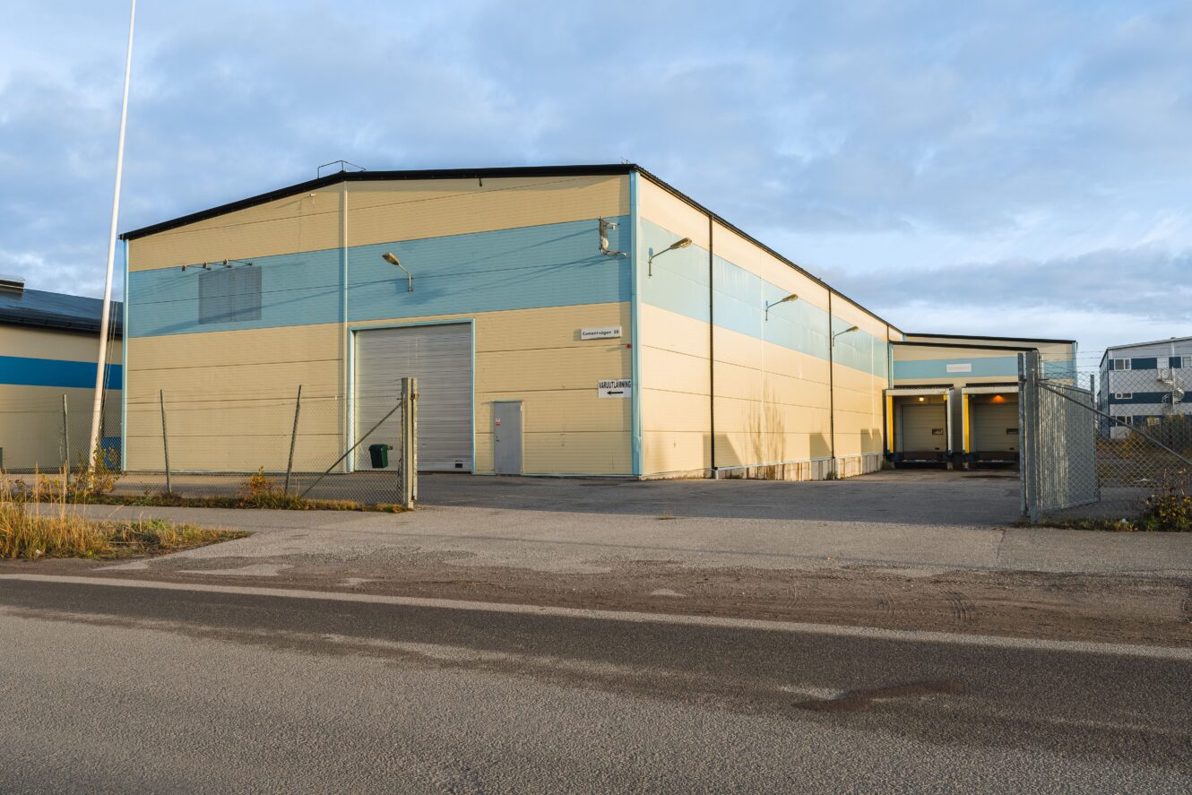 Ett stort gult och blått industrilager med stängsel, lastbryggor och en stängd rullport på en asfalterad tomt.