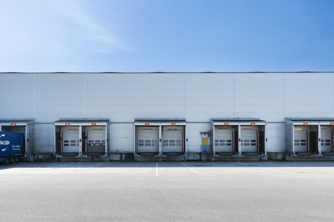 En rad med sex stängda lastkajdörrar vid ett lager, med en tom parkeringsplats framför under en klarblå himmel.