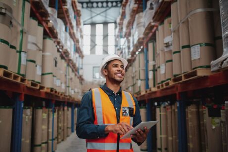 En man i skyddshjälm och skyddsväst står i en lagergång, håller i en surfplatta och tittar upp på staplade pallar med varor.