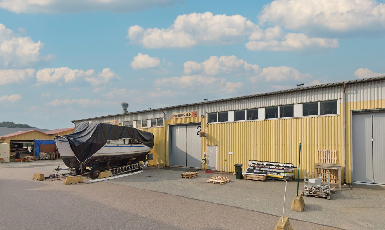 Ett gult industrilager med metallfasad, en delvis täckt båt, träpallar och diverse utrustning utomhus på en asfalterad tomt under en molnig himmel.