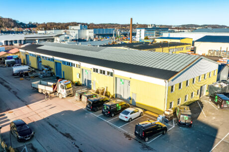 En stor gul industrilagerbyggnad med parkerade bilar och skåpbilar framför, belägen i ett industriområde med fabriker och träd i bakgrunden.