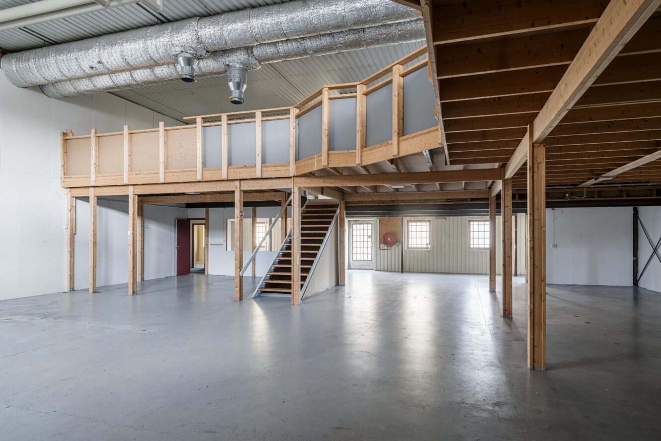 Tomma industrilager med mezzanin i synligt trä, metallkanaler i taket, betonggolv och stora fönster som släpper in naturligt ljus.