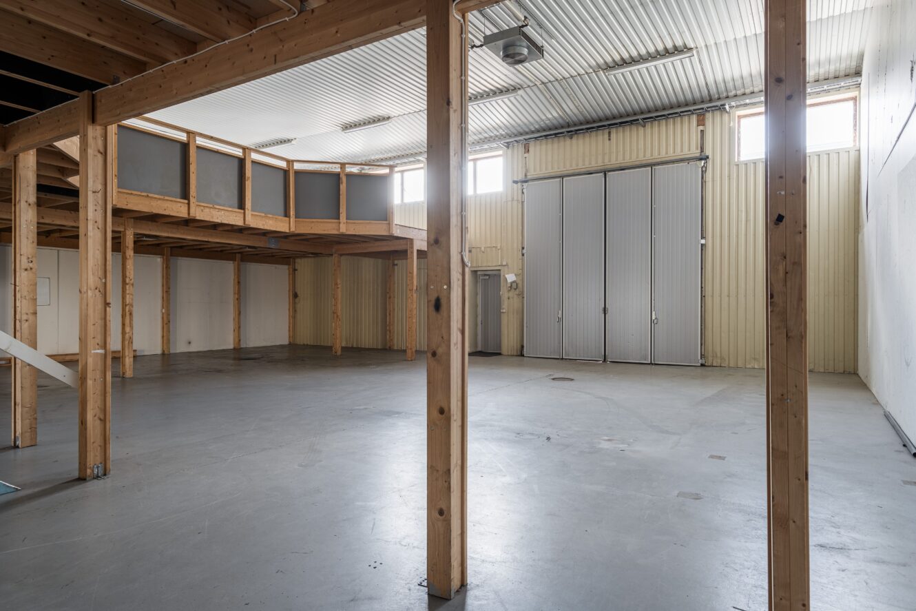 Interiör i en tom industrilokal med högt i tak, synliga träbjälkar, mezzaninplan, betonggolv och stora metalldörrar.