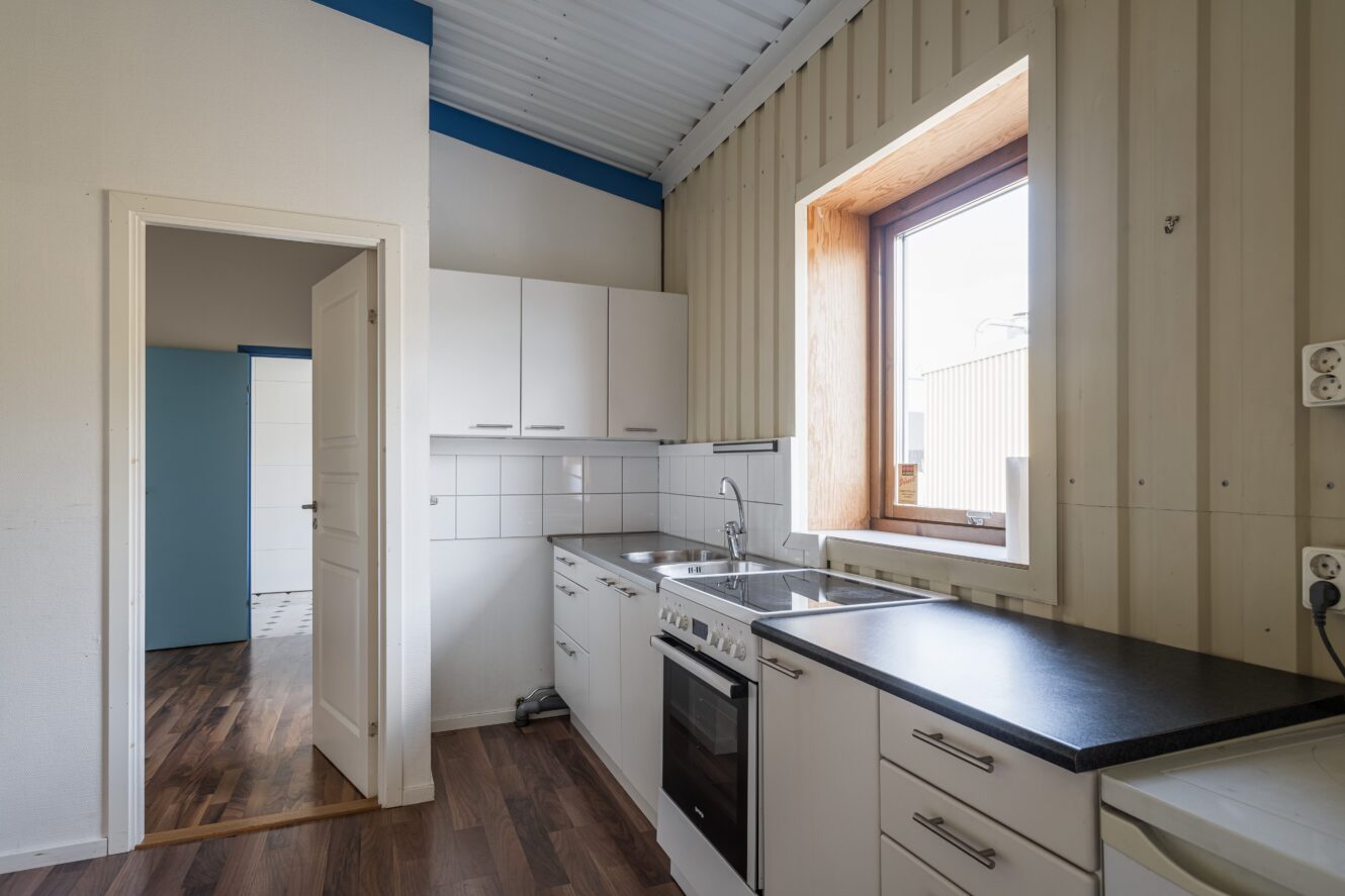 Ett litet, modernt kök med vita skåp, svarta bänkskivor, inbyggd ugn, diskho och ett stort fönster som släpper in naturligt ljus.