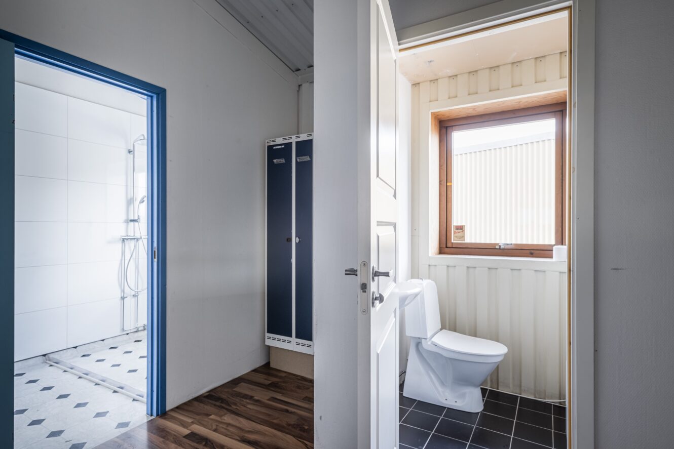 Ett litet badrum med vit toalett, svart klinkergolv och ett fönster, bredvid ett rum med skåp och ett intilliggande duschutrymme.