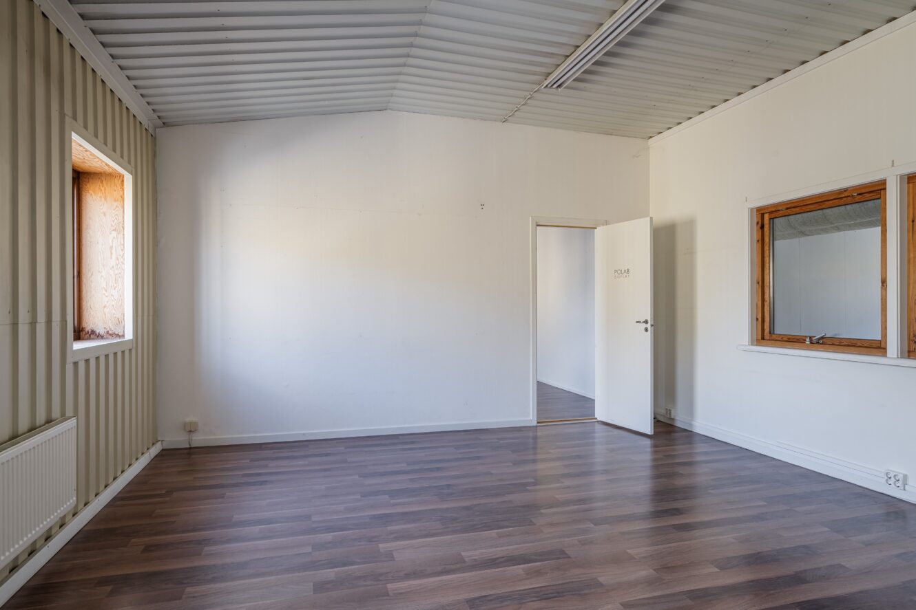 Tomt rum med vita väggar, trämönstrat golv, ett fönster, en öppen dörr till ett annat rum och en stor väggöppning med träram.