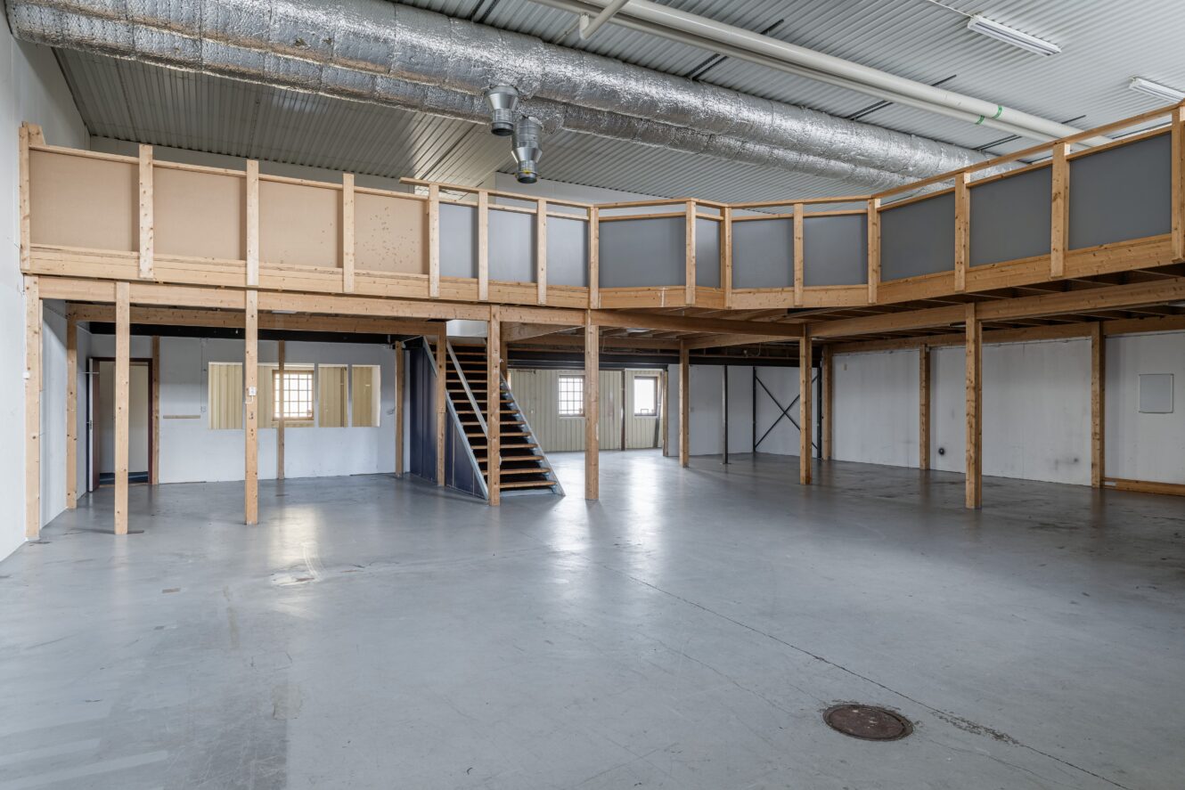 Den rymliga, tomma industriinteriören har ett grått betonggolv, högt tak med synliga kanaler och en mezzanin i trä med trappor.