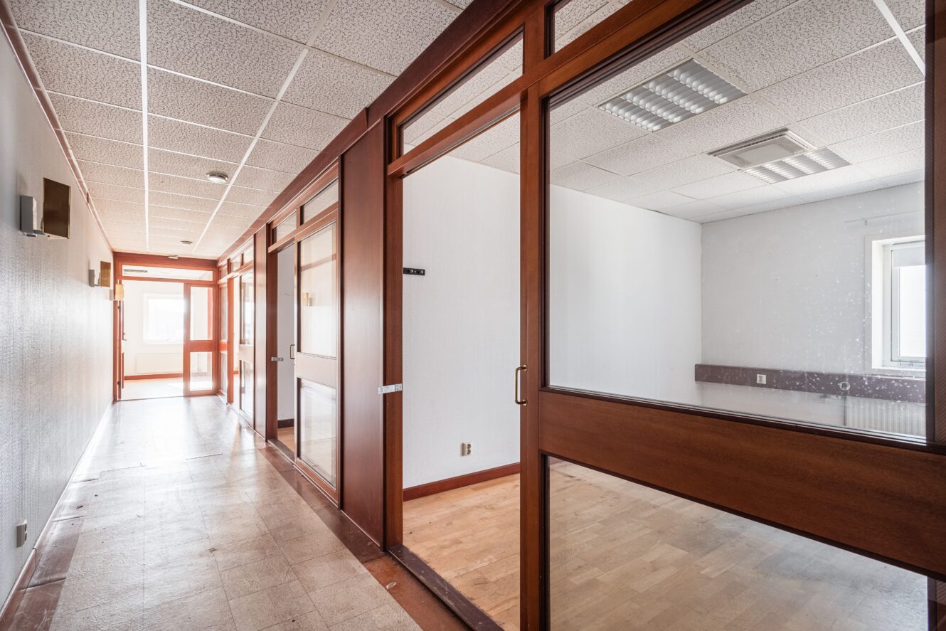 Tom kontorskorridor med rum med glasväggar, träramar, vita väggar och taklampor; naturligt ljus kommer in genom fönster i slutet av korridoren.