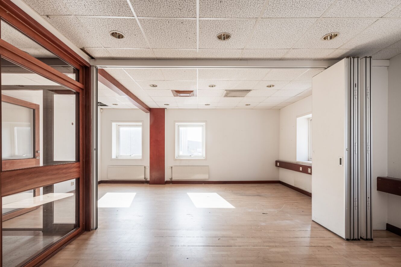 Tomt, väl upplyst rum med trägolv, vita väggar, två fönster, ett tak med infällda lampor och en delvis öppen vikvägg.