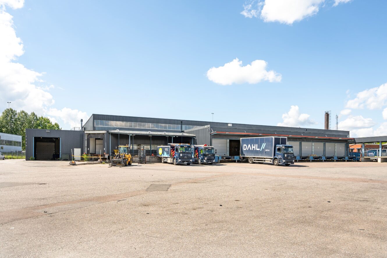 En stor lagerlokal med flera lastkajer, flera lastbilar parkerade och byggutrustning i förgrunden under en delvis molnig himmel.