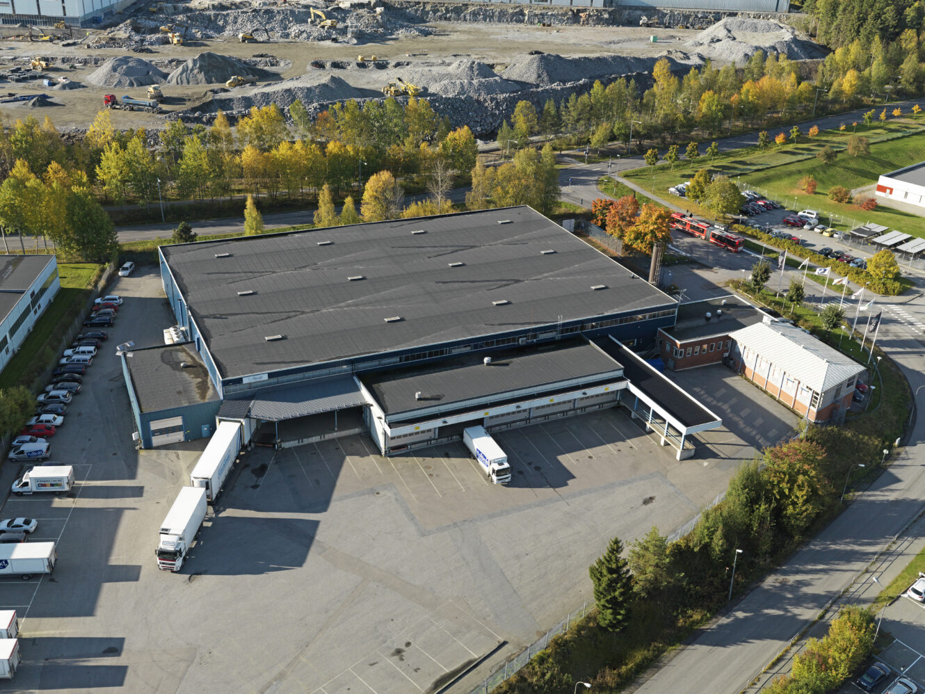 Ett stort industrilager med lastbryggor och parkerade lastbilar, omgivet av träd, bilar och närliggande vägar, sett från ovan.