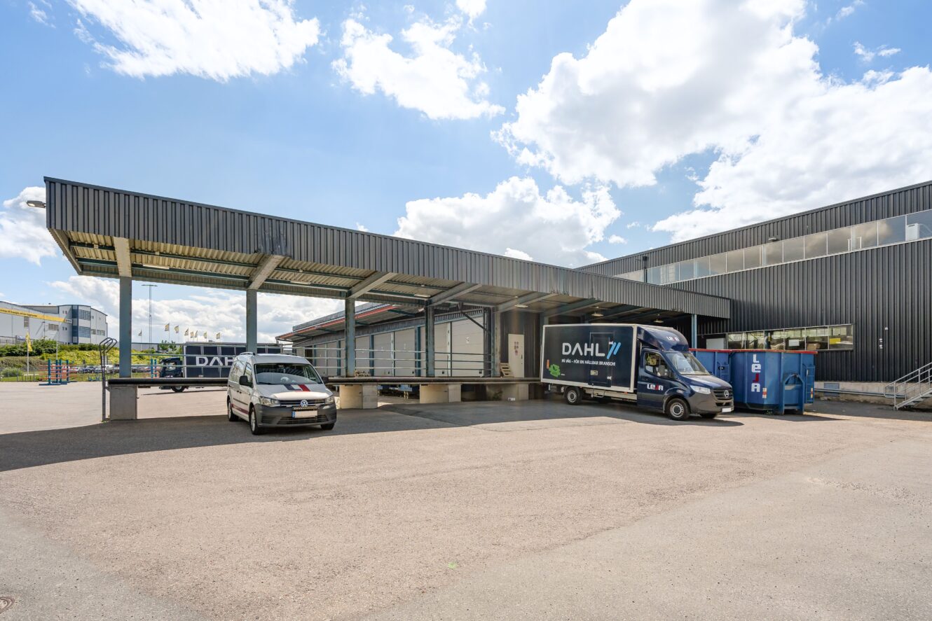 Lastkajsområde med en täckt plattform, en parkerad skåpbil märkt DAHL, en bil och en blå container utanför en stor industribyggnad under en delvis molnig himmel.