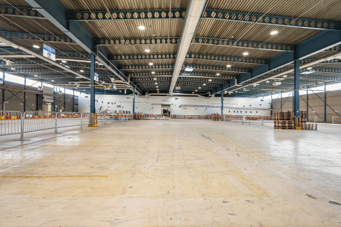 Rymlig, tom industrilagerlokal med högt i tak, synliga bjälkar och utspridda pallar och barriärer längs sidorna.