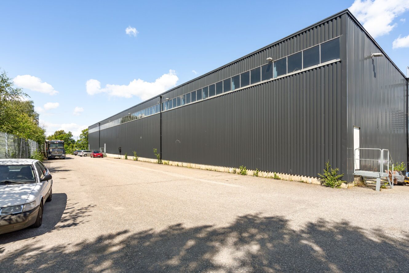 Ett stort, grått industrilager med höga fönster och en asfalterad uppfart; flera parkerade bilar syns längs byggnaden under en klar himmel.