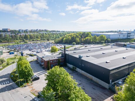 Flygfoto över ett industriellt lager, parkeringsplats fylld med bilar, gröna träd i närheten, under en delvis molnig himmel.