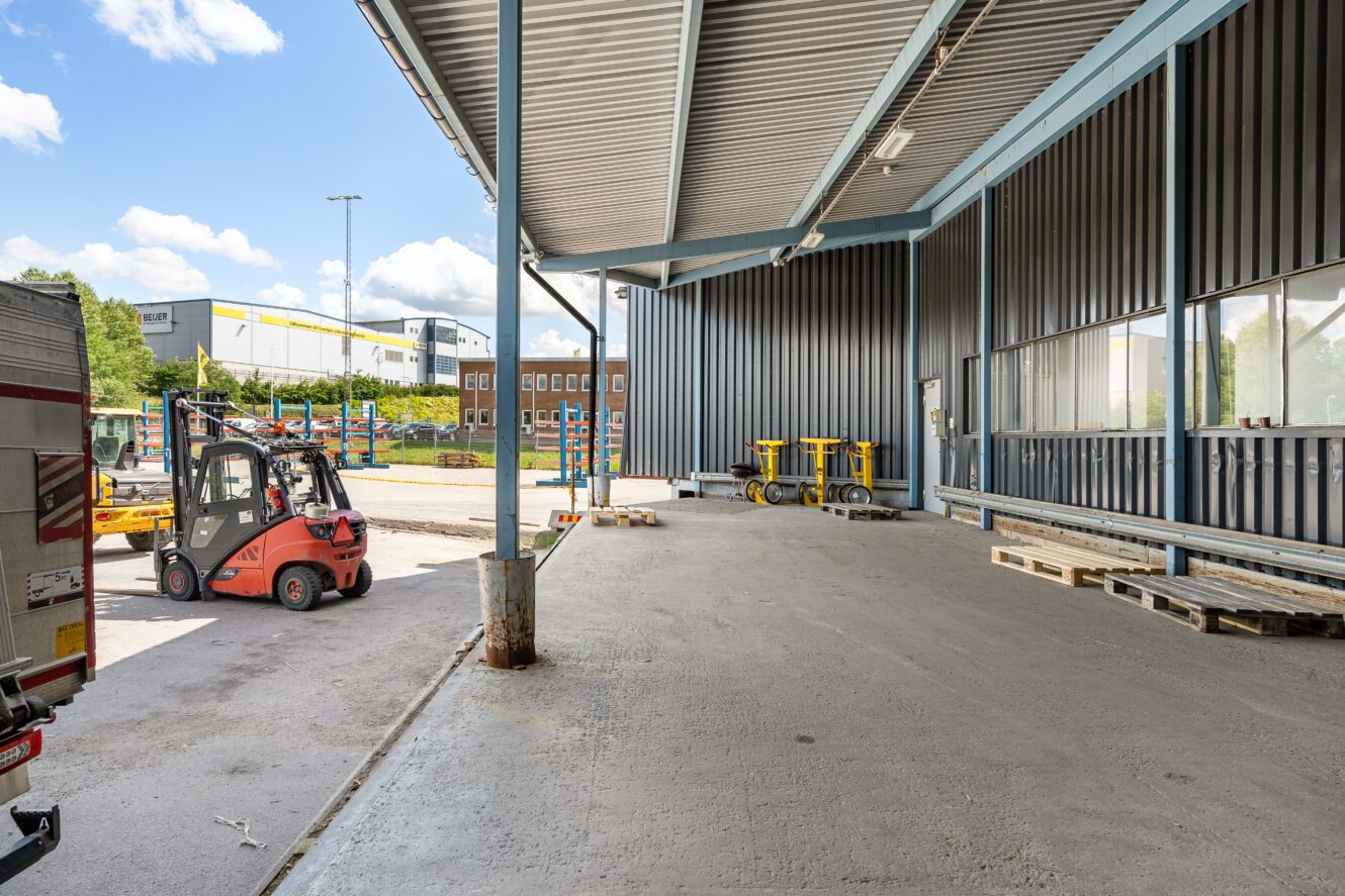 En gaffeltruck kör nära en lastkaj utanför en industribyggnad, där pallar, vagnar och annan lagerutrustning syns.