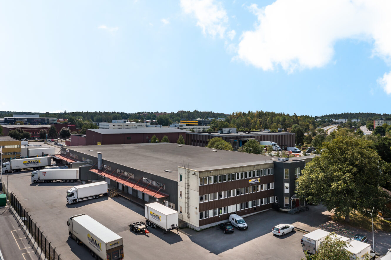 Ett stort kommersiellt lagerkomplex med flera parkerade lastbilar, några bilar och omgivande träd under en klar himmel.