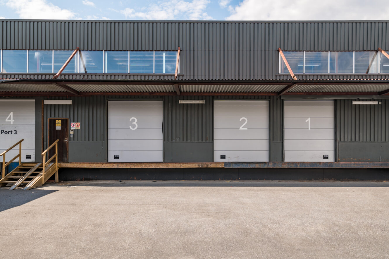 En grå industriell lastkaj med fyra numrerade portar (4, 3, 2, 1) och en metalltrappa som leder till en mörk dörr till vänster.