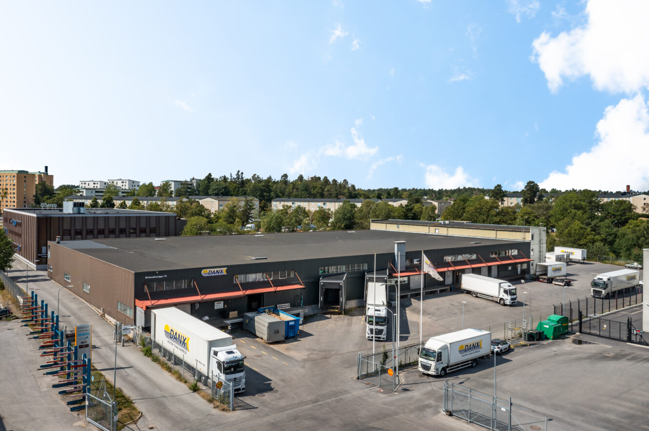 En logistikanläggning med flera lastbilar parkerade utanför, omgiven av ett staket och träd i bakgrunden under en delvis molnig himmel.