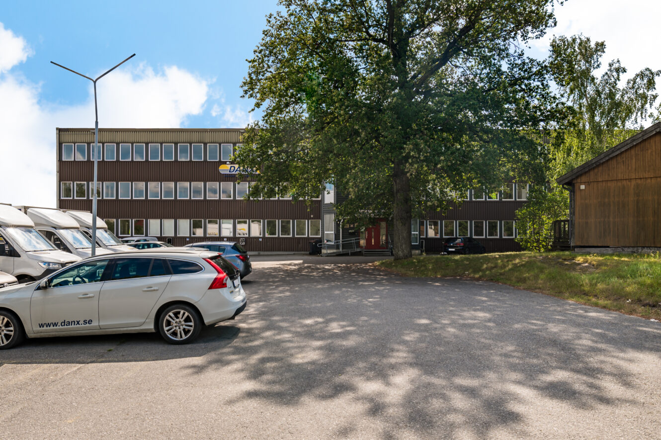 En kontorsbyggnad i tre våningar med DANX-logotypen, omgiven av träd, parkerade bilar och vita skåpbilar i förgrunden.