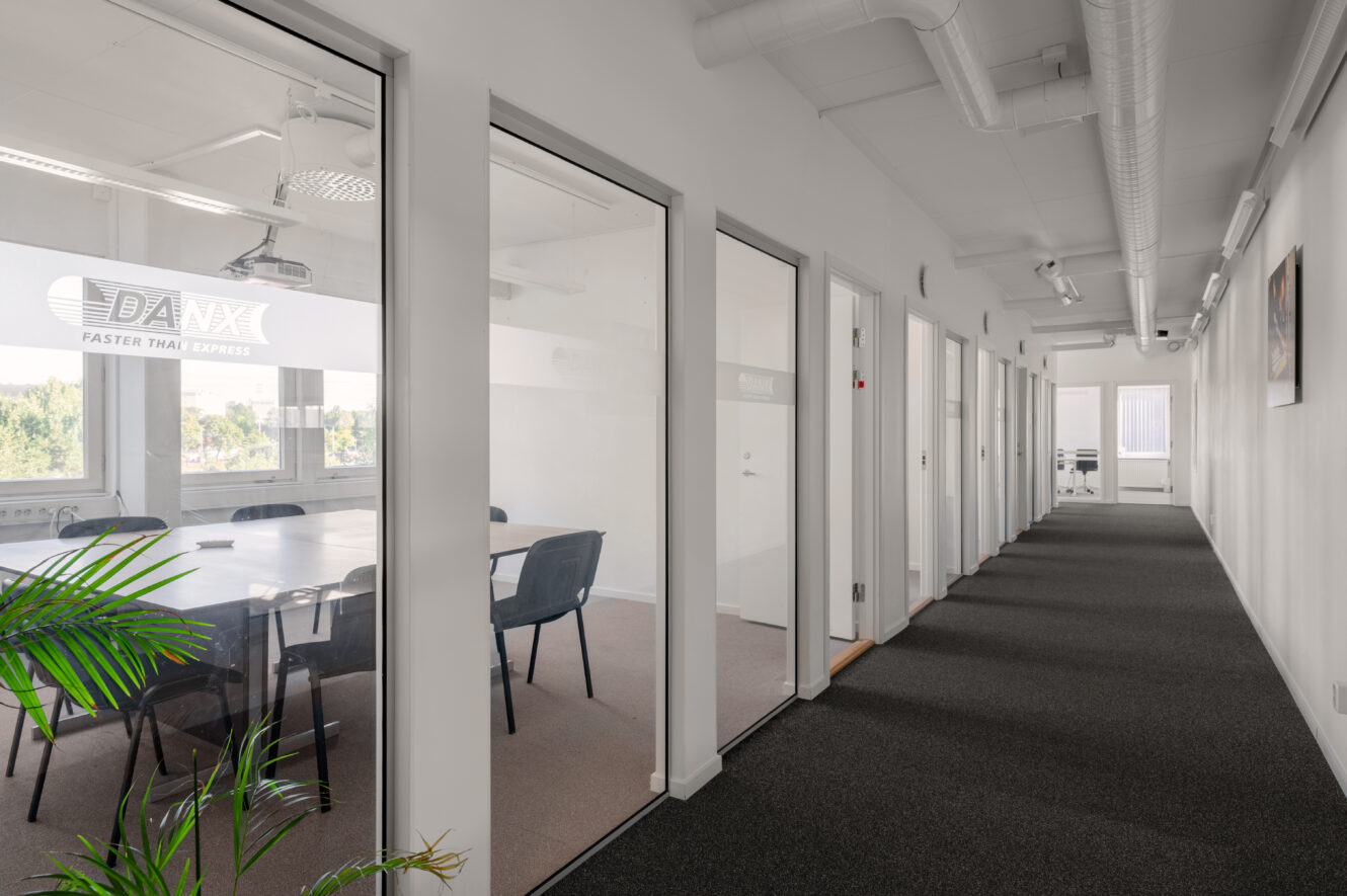 En modern kontorskorridor med mötesrum med glasfasad till vänster, stängda dörrar till höger och naturligt ljusinsläpp genom fönster i båda ändar.