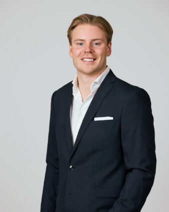 En ung man med blont hår, klädd i mörk kostym och vit skjorta, står mot en ljus bakgrund och ler mot kameran.