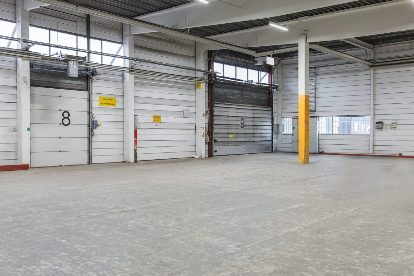 Tom lagerlokal med betonggolv, vita metallväggar, högt i tak och numrerade dörrar till lastkajen.