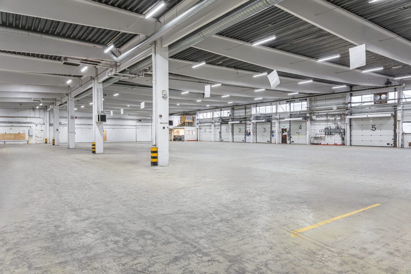 Rymlig, tom industrilagerinredning med betonggolv, vita väggar, numrerade lastkajsdörrar och takbelysning.