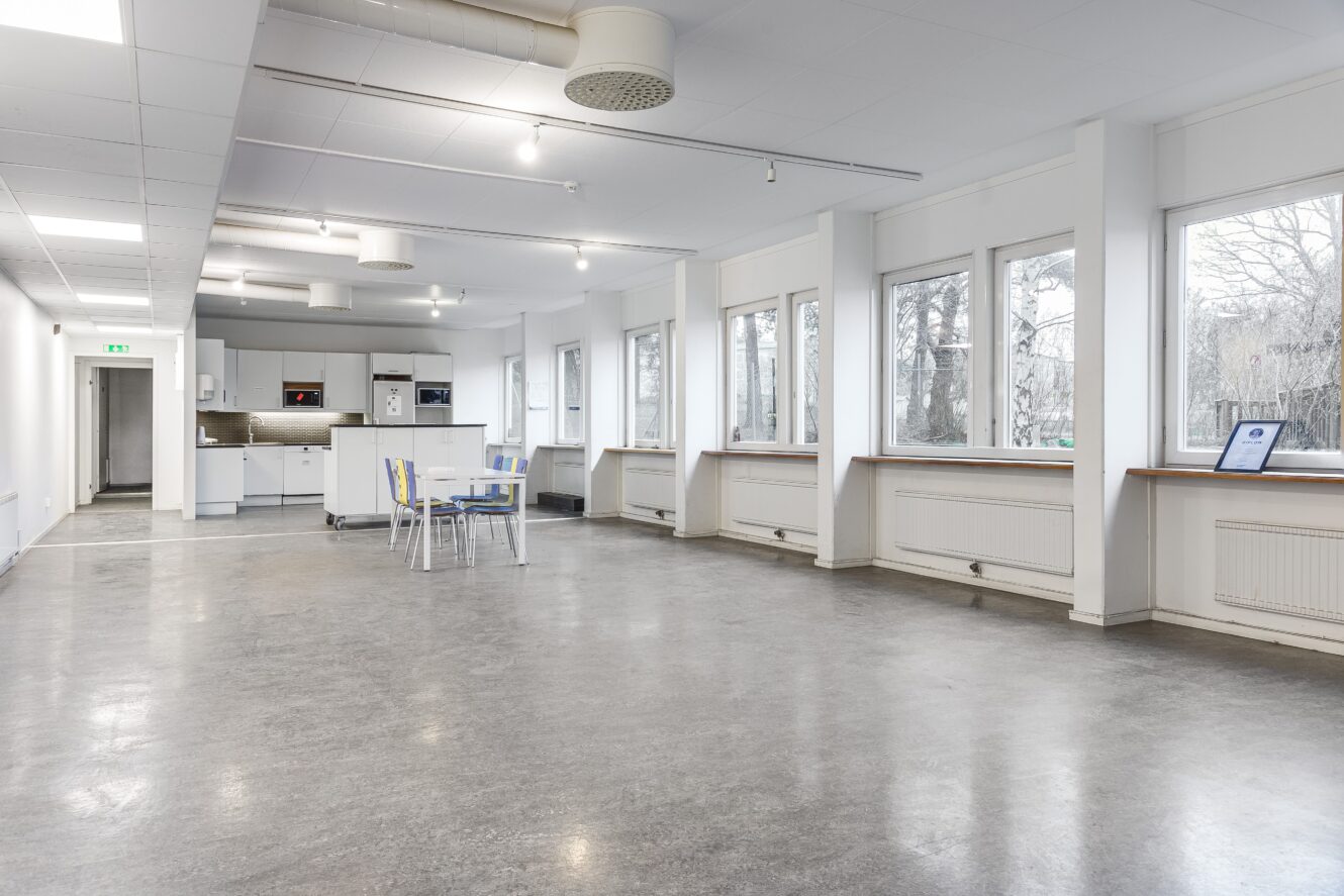 Ett rymligt, modernt rum med ett pentry, ett matbord med fyra stolar, stora fönster och grått golv. Utrymmet ser ljust och minimalistiskt ut.